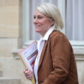 Smiling woman with short blonde hair in a brown jacket, holding folders and a pen outdoors beside a stone building.
