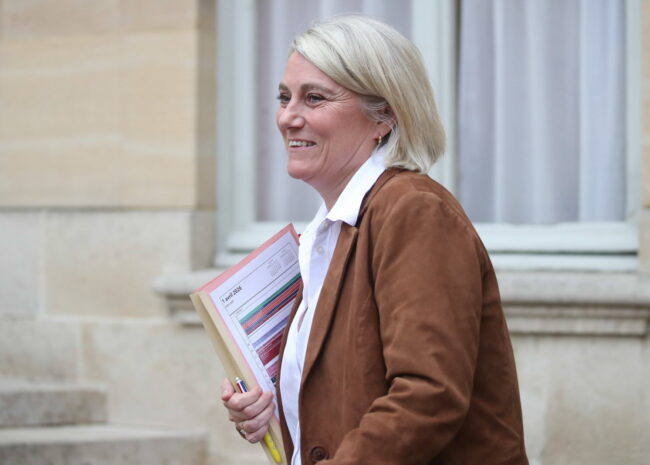Smiling woman with short blonde hair in a brown jacket, holding folders and a pen outdoors beside a stone building.