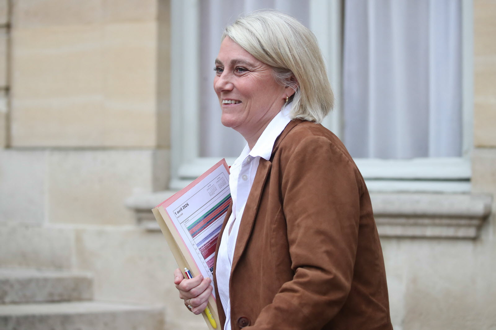 Smiling woman with short blonde hair in a brown jacket, holding folders and a pen outdoors beside a stone building.
